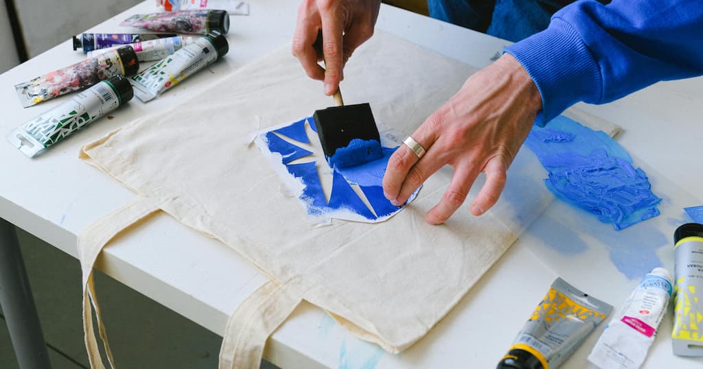Artist uses stencil to apply blue paint to a canvas bag in a creative studio environment